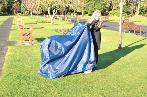 Person unfolding a large blue storage cover over a mobility scooter in a sunny park