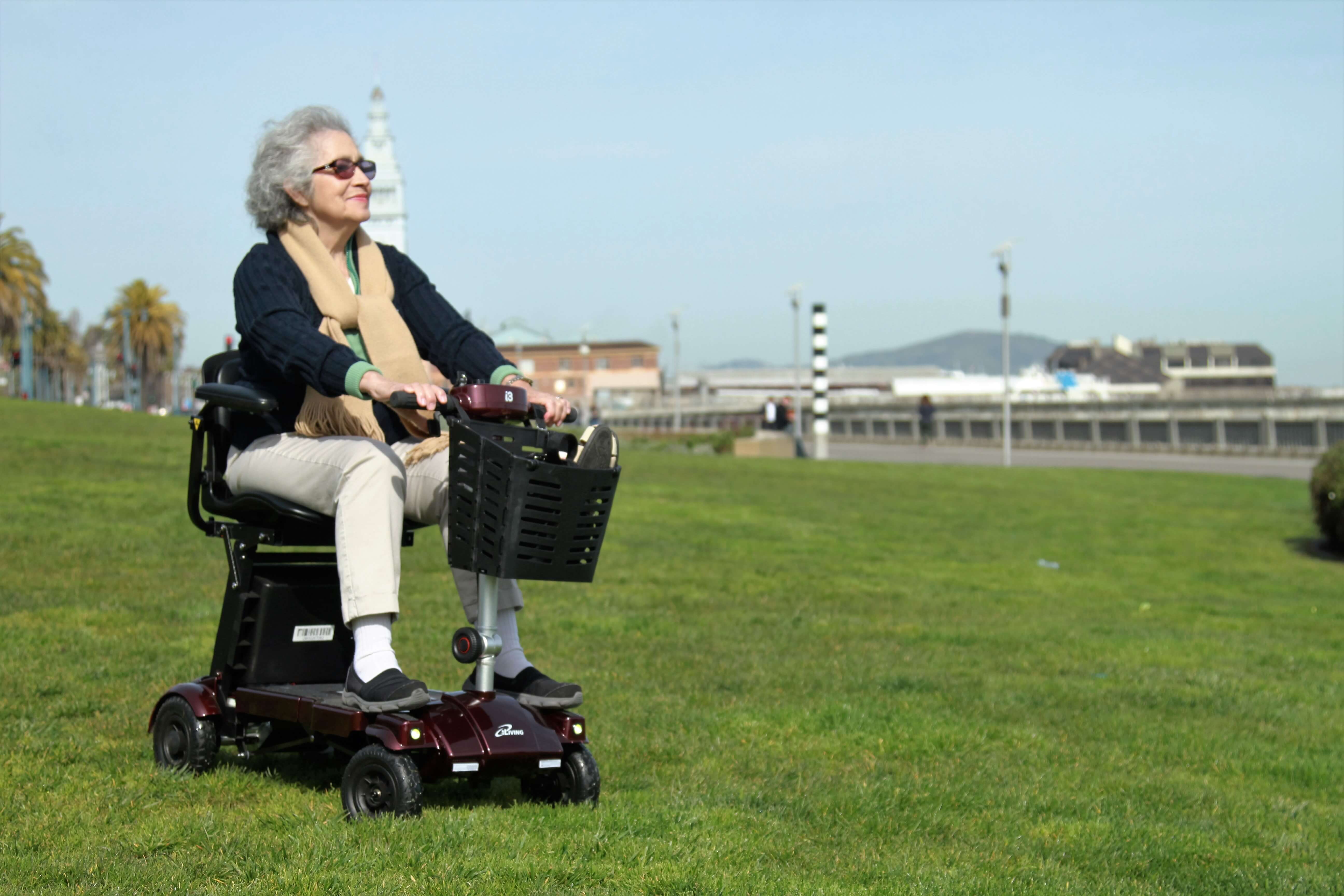 Senior woman with sunglasses enjoying a ride on a portable mobility scooter across a grassy field.