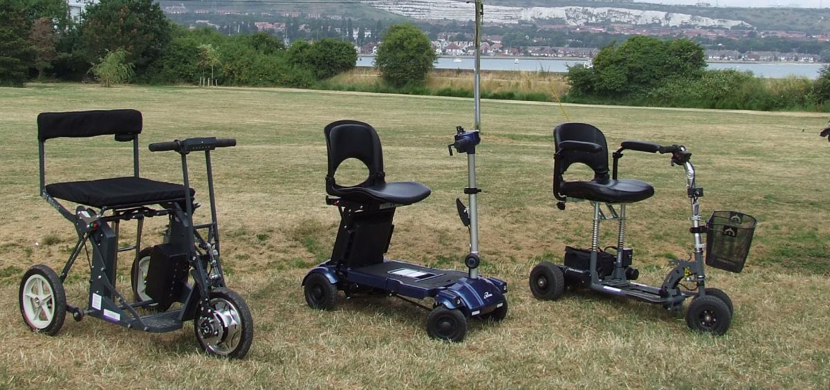 Three different models of lightweight mobility scooters displayed side by side on a grassy field, with a scenic waterfront backdrop.