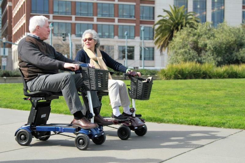 Two seniors enjoying a ride on i3 mobility scooters in an urban park, with a lush green and urban backdrop.