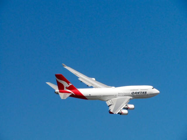 A large passenger jet with a red and white livery, identifiable as a Qantas airliner, is captured mid-flight against a clear blue sky, with its landing gear retracted, suggesting it is cruising rather than preparing to land.