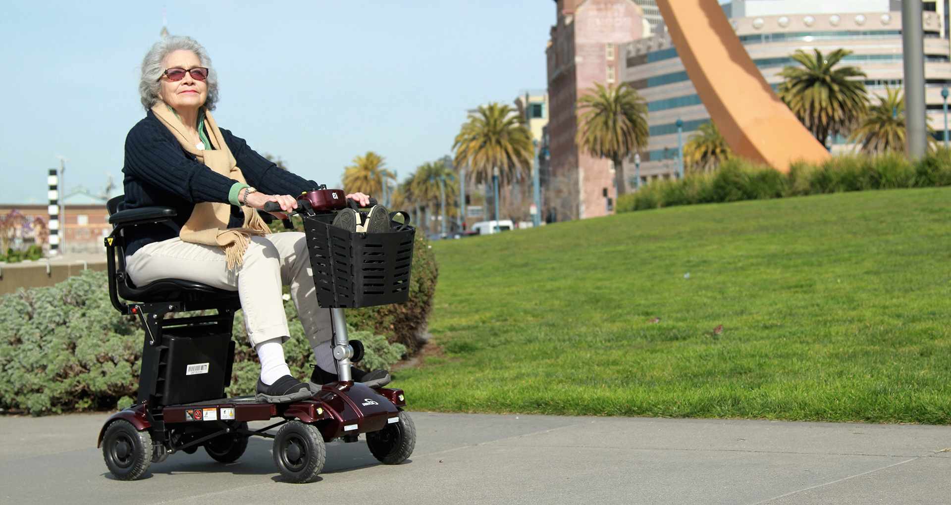 An elderly lady riding a portable i3 mobility scooter through a park on a sunny day, enjoying the ride.